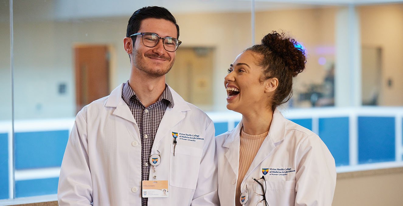 two SOM students stand next to each other, one smiling, one laughing, in the atrium of the Rowan Medicine Building in Sewell