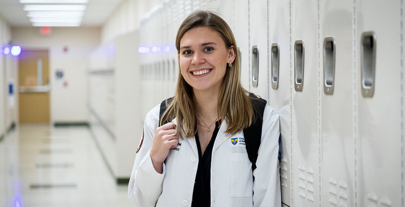 An SOM student smiles at the camera while standing in the hallway in front of lockers