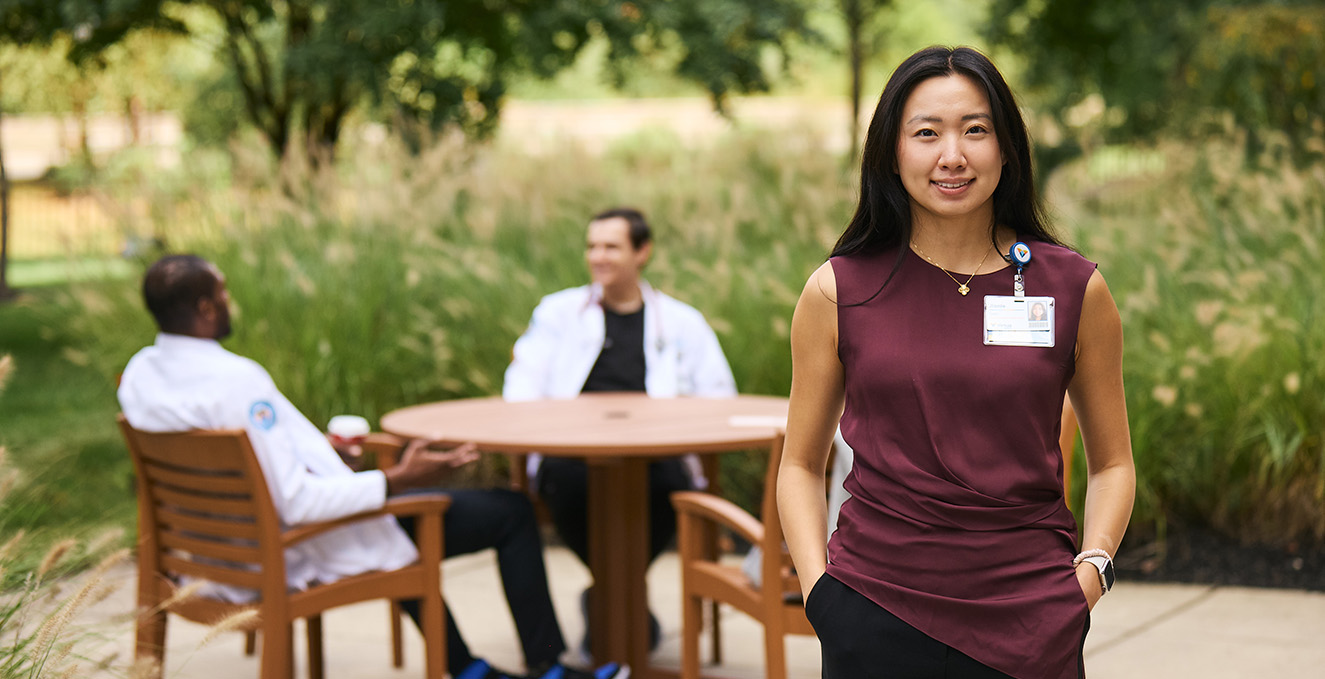 a smiling SOM student stands outside Virtua hospital while fellow medical students sit at a table in the background