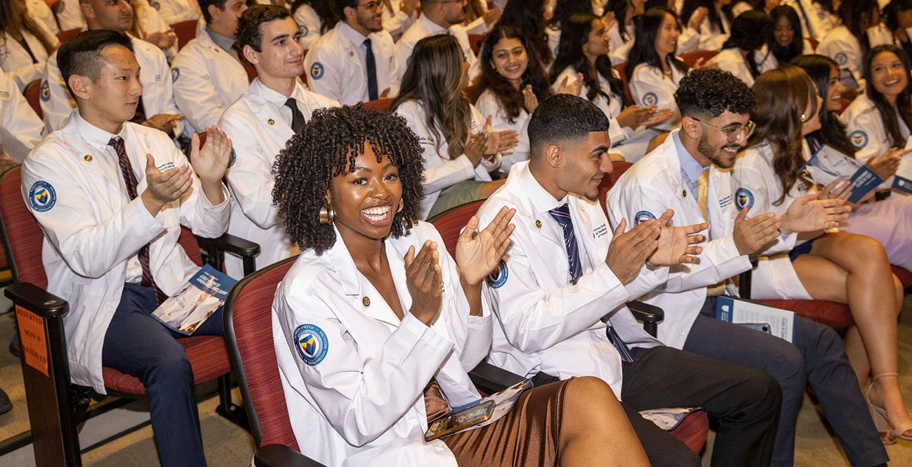 One SOM student smiles at the camera while sitting in the audience filled with other SOM students during the white coat ceremony