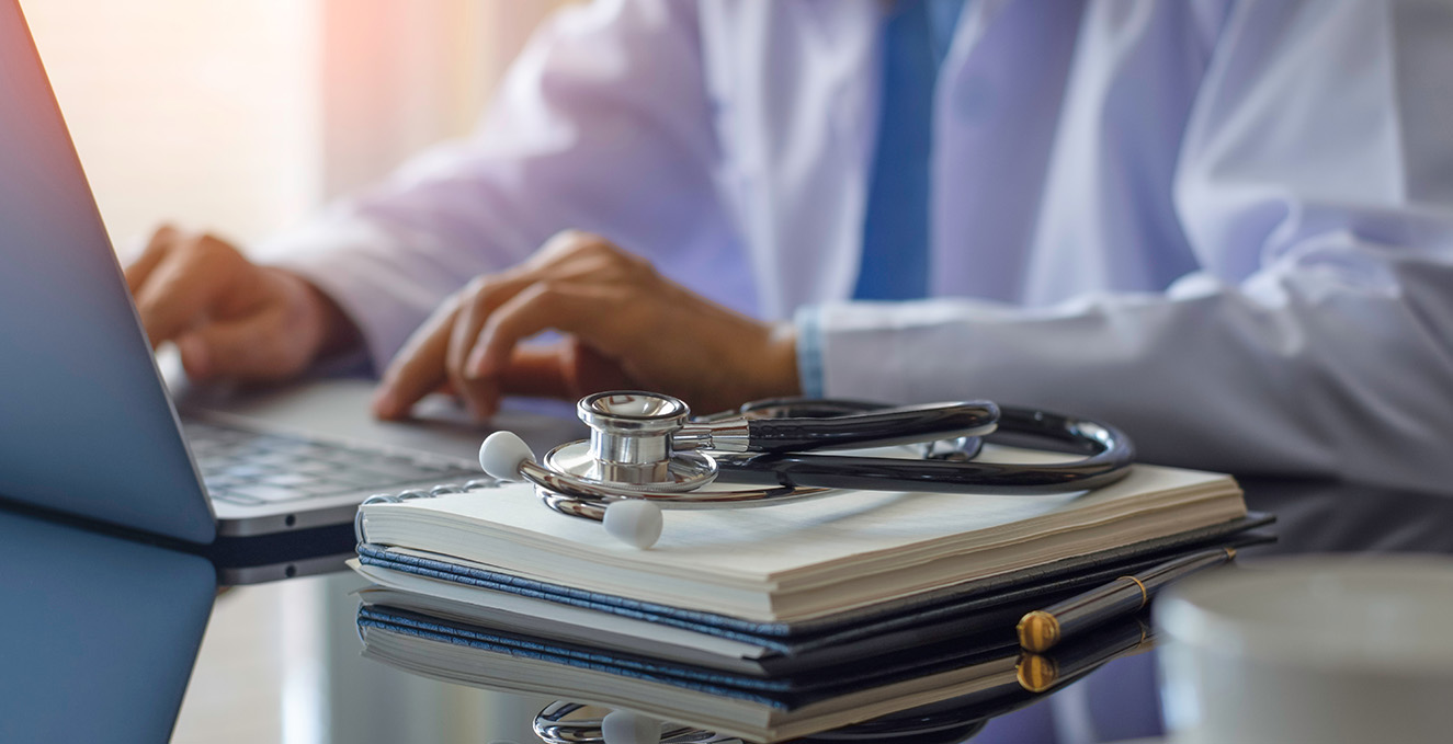 hands working at a desk laptop, with a stethoscope and book