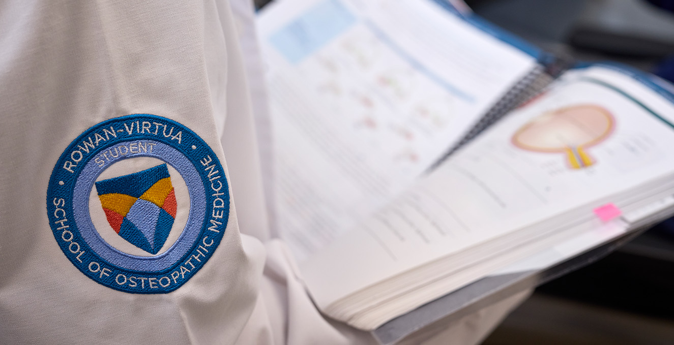 A Rowan-Virtua SOM student patch is visible on the sleeve of a white coat while a student reads a text book