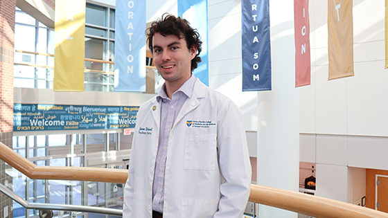 SOM medical student Jason Richwall wears his white coat and smiles while standing on the landing of the staircase in the academic center. Banners hang on the walls behind him 