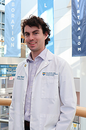 medical student Jason Richwall smiles while wearing his white coat and standing on the landing in the SOM academic center with banners hanging behind him