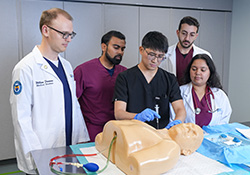 a medical student works on a procedure with a syringe on a mannequin while other med students watch