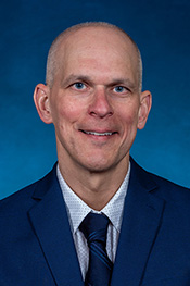 Curt Cackovic smiles for the camera while wearing a blue jacket, blue tie and a white patterned shirt