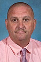 Allen Ecker smiles for the camera while wearing a pick shirt and a purple tie