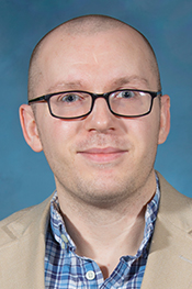 Daniel Hurst smiles for the camera while wearing glasses, a plaid shirt and tan jacket