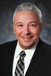 Dean Micciche smiles at the camera while wearing a white button down shirt, black jacket and a black striped tie