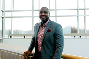 John Nwafor smiles into the camera while standing in the atrium of the Academic Center on the Stratford Campus