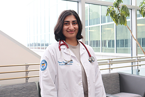 Rowan-Virtua SOM medical student smiles while wearing her white coat and standing in front of windows in the ROwan Medicine building.