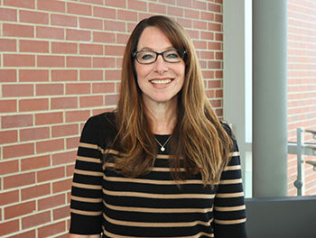 Tara Shontz, wearing a black and tan striped sweater stands in an SOM building with a brick wall and column behind her