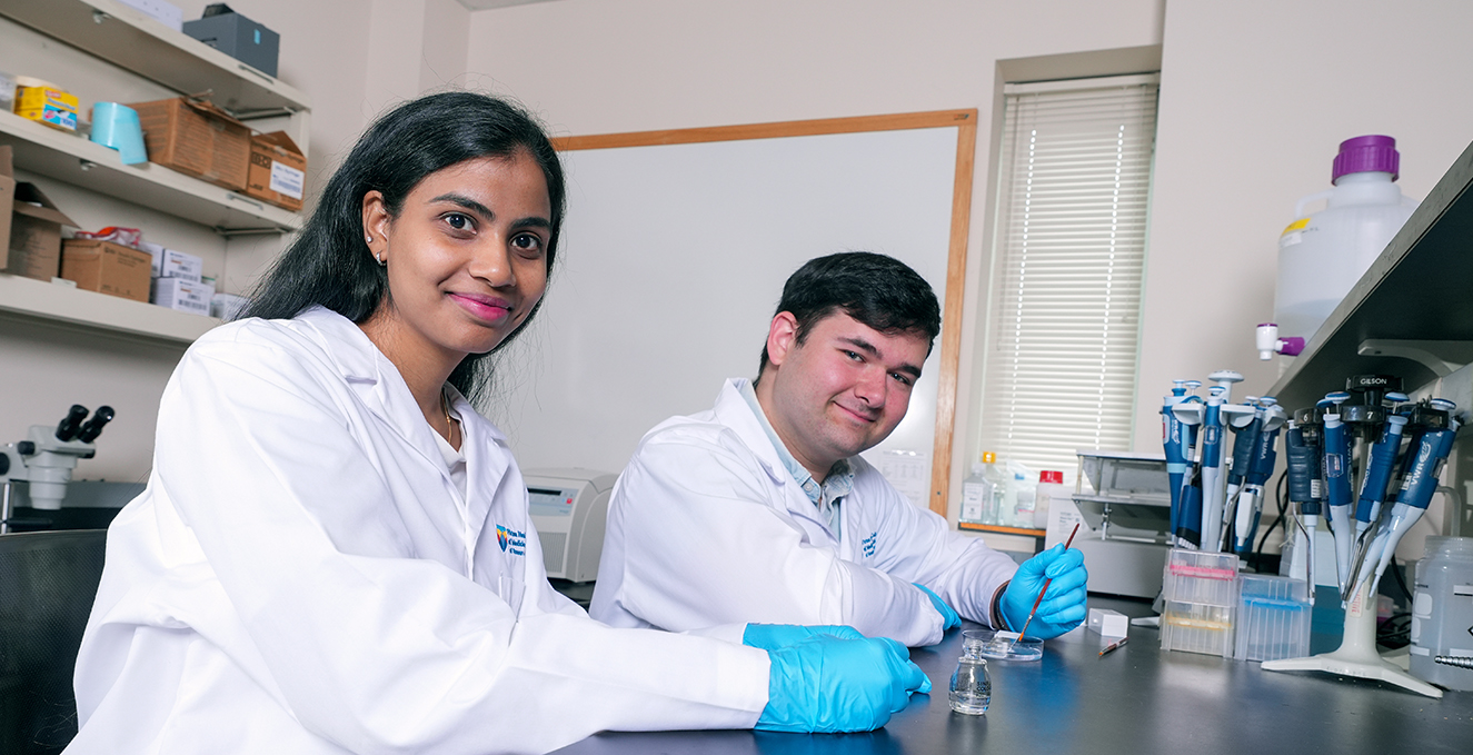 two research students smile at the camera while working at a lab table
