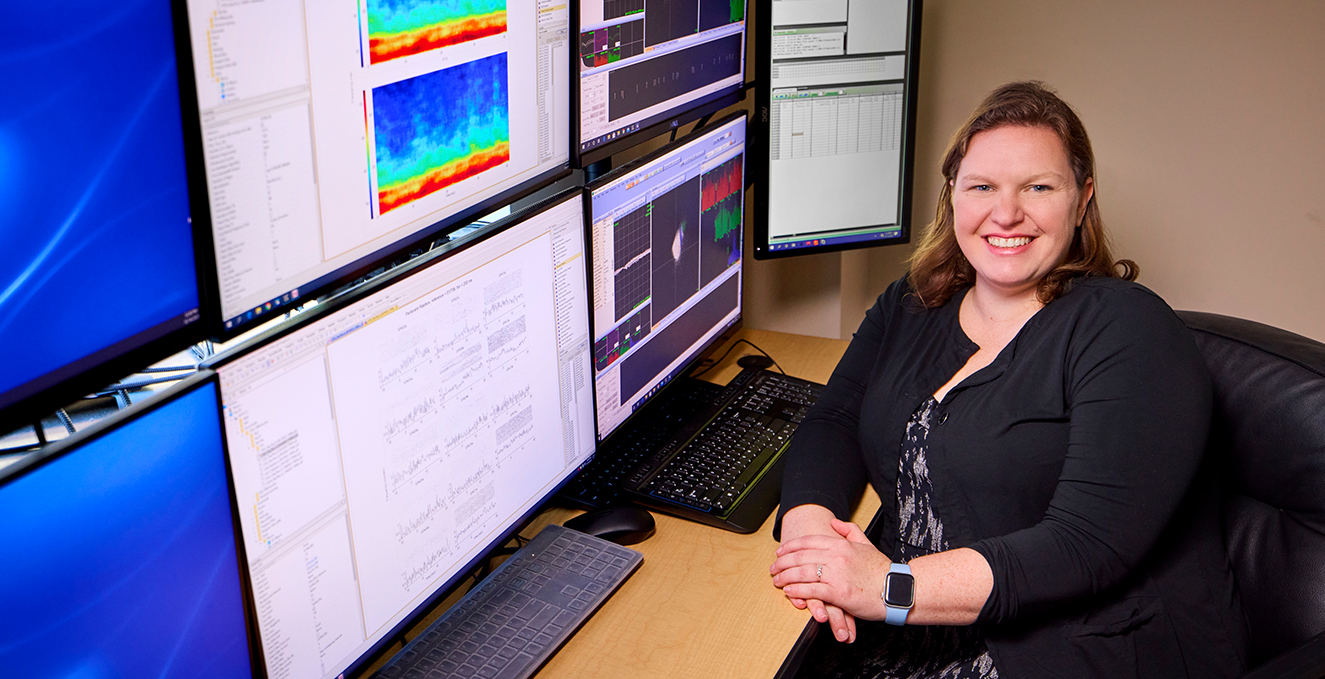 a research faculty member smiles at the camera while sitting in front of data analyzation on monitors