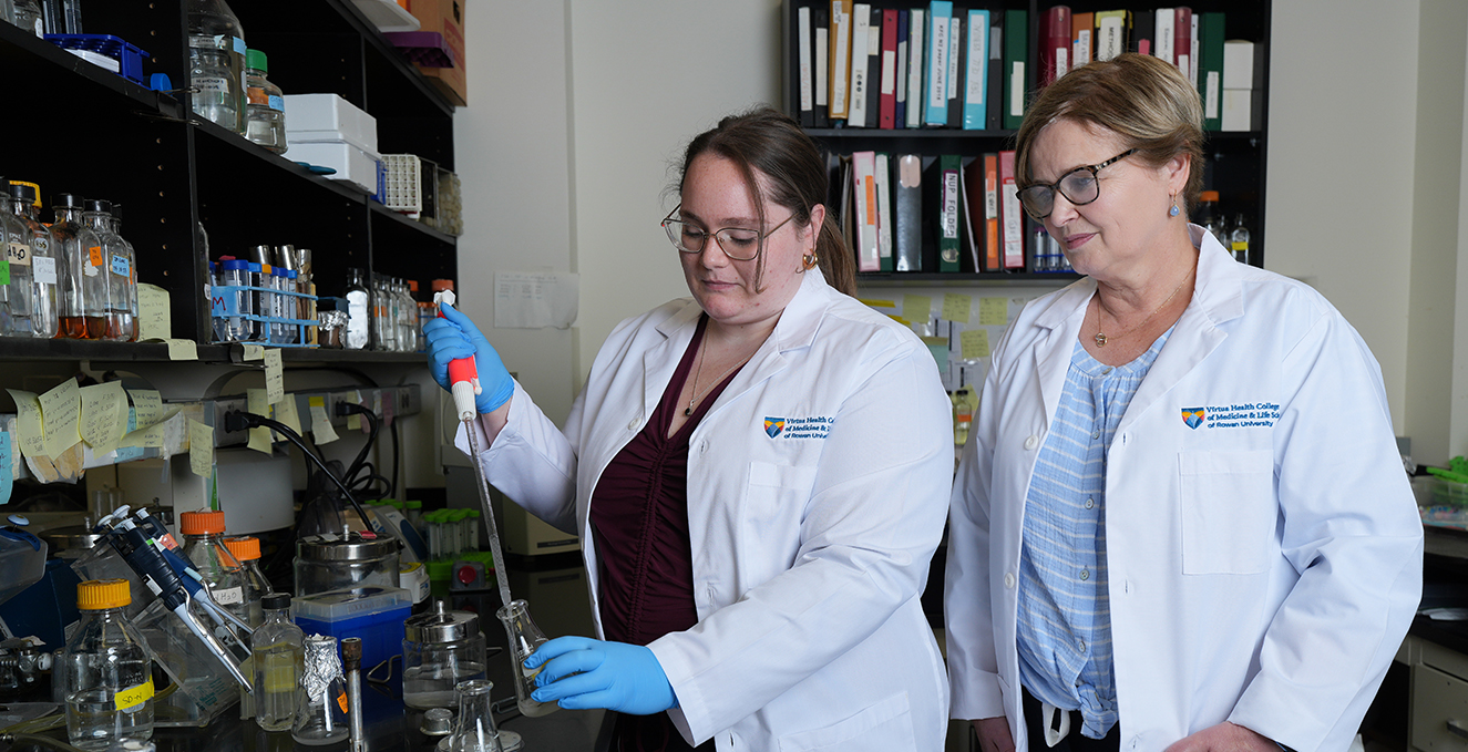 a research student puts liquid in a beaker while a faculty researcher looks at her in the lab