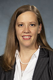 Katherine Milani smiles for the camera while wearing a dark, pinstripe suit jacket, a white shirt and a pearl necklace