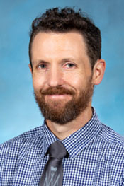 Jeffrey Powers smiles at the camera while wearing a blue checked shirt and a grey tie