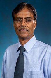 Lakshman Segar smiles at the camera while wearing his glasses, a blue button down shirt and a blue tie